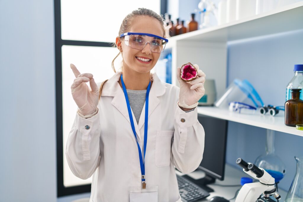 Young woman working at scientist laboratory holding geode smiling happy pointing with hand and finger to the side